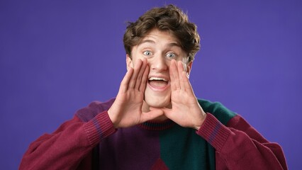 Portrait of happy smiling young man 20s yelling hey with hands up to mouth isolated on purple background. Studio real emotions.