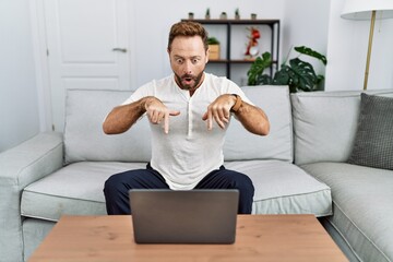 Poster - Middle age man using laptop at home pointing down with fingers showing advertisement, surprised face and open mouth