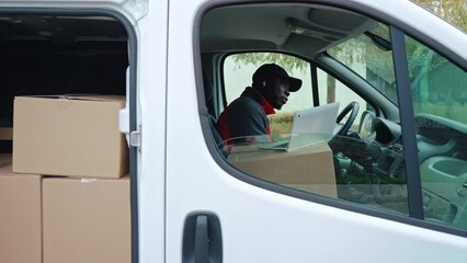 Wall Mural - Black young adult delivery man sitting in white van with cardboard box packages filling out documents and working on laptop. . High quality 4k footage