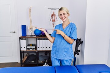 Canvas Print - Young physiotherapist woman working at pain recovery clinic amazed and smiling to the camera while presenting with hand and pointing with finger.