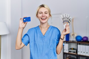 Canvas Print - Young physiotherapist woman holding credit card and dollars banknotes smiling and laughing hard out loud because funny crazy joke.