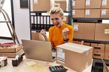 Young blonde woman working at small business ecommerce using laptop screaming proud, celebrating victory and success very excited with raised arms