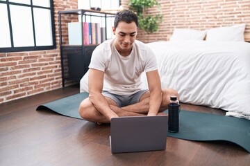 Wall Mural - Young hispanic man using laptop sitting on yoga mat at bedroom