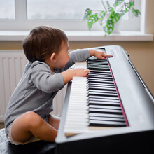 Toddler Baby Plays Piano In Home Living Room, Music Lessons. Happy Child Boy Learns To Play Music On An Electric Piano. Kid Boy Age One Year Four Months