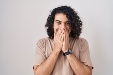 Sticker - Hispanic man with curly hair standing over white background laughing and embarrassed giggle covering mouth with hands, gossip and scandal concept