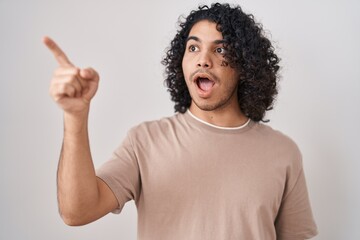 Poster - Hispanic man with curly hair standing over white background pointing with finger surprised ahead, open mouth amazed expression, something on the front