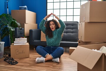 Wall Mural - Young hispanic woman sitting on the floor at new home in shock face, looking skeptical and sarcastic, surprised with open mouth