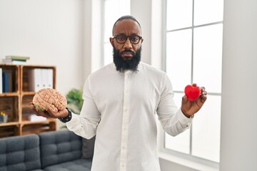 Canvas Print - African american man working at therapy office holding brain and heart relaxed with serious expression on face. simple and natural looking at the camera.