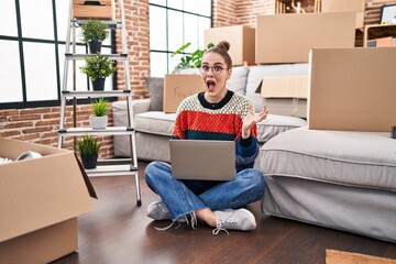 Poster - Young hispanic girl sitting on the floor at new home with laptop celebrating victory with happy smile and winner expression with raised hands