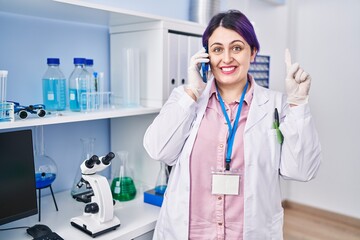 Sticker - Plus size woman wit purple hair working at scientist laboratory speaking on g the phone smiling with an idea or question pointing finger with happy face, number one