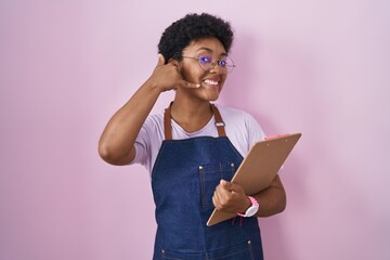 Poster - Young african american woman wearing professional waitress apron holding clipboard smiling doing phone gesture with hand and fingers like talking on the telephone. communicating concepts.