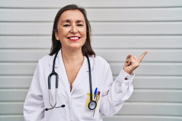 Poster - Middle age hispanic woman wearing doctor uniform and stethoscope smiling happy pointing with hand and finger to the side