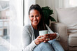 © Westend61 - Happy mature woman sitting with coffee cup by window at home