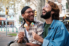 Happy Gay Couple Enjoying Candy Sitting On Bench