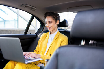 Beautiful young woman with short hair and colorful business suit remote working with laptop in a car - Corporate businesswoman on a business travel