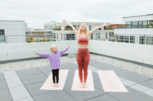 Daughter With Arms Outstretched Looking At Mother Practicing Yoga On Rooftop