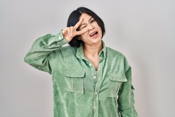 Poster - Young asian woman standing over white background doing peace symbol with fingers over face, smiling cheerful showing victory