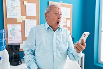 Canvas Print - Senior man with grey hair working at the office doing video call with smartphone scared and amazed with open mouth for surprise, disbelief face