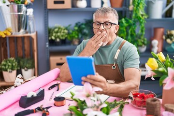 Wall Mural - Hispanic man with grey hair working at florist shop doing video call with tablet covering mouth with hand, shocked and afraid for mistake. surprised expression