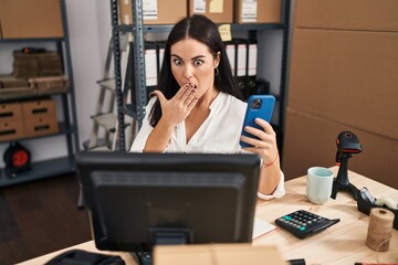 Wall Mural - Young hispanic woman working at small business ecommerce with smartphone covering mouth with hand, shocked and afraid for mistake. surprised expression