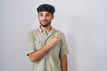 Sticker - Arab man with beard standing over white background pointing with hand finger to the side showing advertisement, serious and calm face