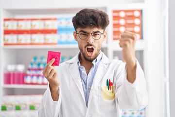 Wall Mural - Arab man with beard working at pharmacy drugstore holding condom afraid and shocked with surprise and amazed expression, fear and excited face.