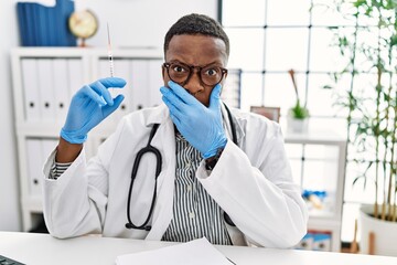 Canvas Print - Young african doctor man holding syringe at the hospital shocked covering mouth with hands for mistake. secret concept.