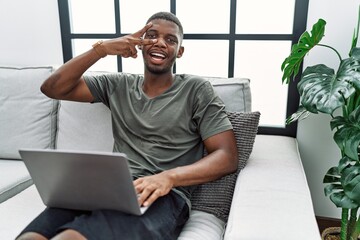 Canvas Print - Young african american man using laptop at home sitting on the sofa doing peace symbol with fingers over face, smiling cheerful showing victory