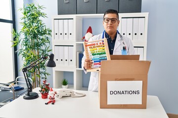 Sticker - Hispanic young doctor man donating toys at the clinic relaxed with serious expression on face. simple and natural looking at the camera.