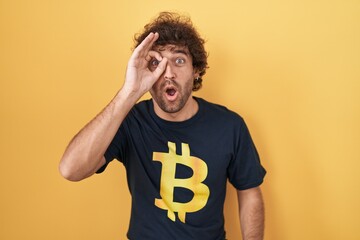 Sticker - Hispanic young man wearing bitcoin t shirt doing ok gesture shocked with surprised face, eye looking through fingers. unbelieving expression.