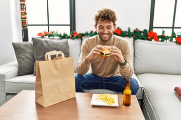 Canvas Print - Young hispanic man eating take away hamburger sitting by christmas decor at home