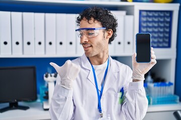 Poster - Hispanic man working at scientist laboratory showing smartphone screen pointing thumb up to the side smiling happy with open mouth