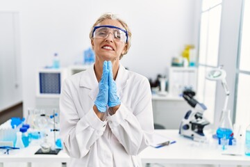 Wall Mural - Middle age blonde woman working at scientist laboratory begging and praying with hands together with hope expression on face very emotional and worried. begging.