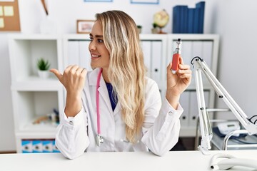 Wall Mural - Young beautiful doctor woman holding electronic cigarette at the clinic pointing thumb up to the side smiling happy with open mouth