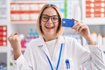 Canvas Print - Young caucasian woman working at pharmacy drugstore holding credit card screaming proud, celebrating victory and success very excited with raised arm