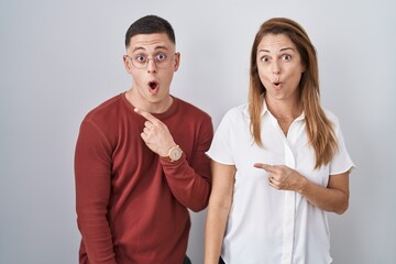 Wall Mural - Mother and son standing together over isolated background surprised pointing with finger to the side, open mouth amazed expression.