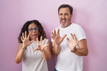 Poster - Middle age hispanic couple together over pink background afraid and terrified with fear expression stop gesture with hands, shouting in shock. panic concept.