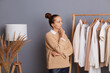 © sementsova321 - Indoor shot of pensive thoughtful woman with bun hairstyle standing in fashion store in front of hangers with attires, thinking choosing clothes to buy.
