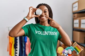 Wall Mural - Young african american woman wearing volunteer uniform holding canned food over eye at charity center