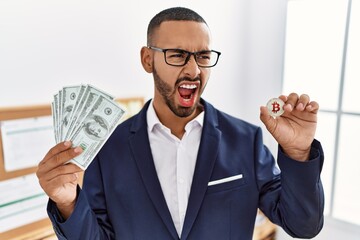 Canvas Print - African american young man holding bitcoin virtual money and dollars banknotes angry and mad screaming frustrated and furious, shouting with anger. rage and aggressive concept.