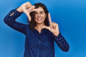 Poster - Young brunette woman with curly hair wearing casual clothes over blue background smiling making frame with hands and fingers with happy face. creativity and photography concept.