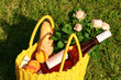 Yellow wicker bag with roses, peaches, baguette and wine on green grass outdoors