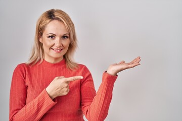 Canvas Print - Blonde woman standing over isolated background amazed and smiling to the camera while presenting with hand and pointing with finger.