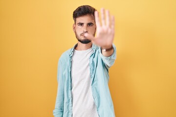 Wall Mural - Young hispanic man with tattoos standing over yellow background doing stop sing with palm of the hand. warning expression with negative and serious gesture on the face.