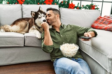 Sticker - Young hispanic man watching movie sitting on sofa with dog by christmas decor at home