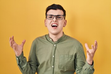 Sticker - Non binary person standing over yellow background crazy and mad shouting and yelling with aggressive expression and arms raised. frustration concept.