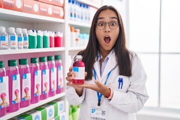 Canvas Print - Young hispanic woman working at pharmacy drugstore holding syrup afraid and shocked with surprise and amazed expression, fear and excited face.