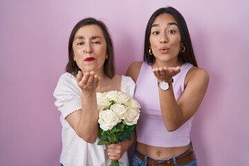 Wall Mural - Hispanic mother and daughter holding bouquet of white flowers looking at the camera blowing a kiss with hand on air being lovely and sexy. love expression.