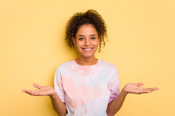 Young Brazilian curly hair cute woman isolated on yellow background makes scale with arms, feels happy and confident.