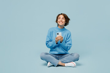 Full body fun young woman wear knitted sweater hold in hand use mobile cell phone look aside on workspace isolated on plain pastel light blue cyan background studio portrait. People lifestyle concept.
